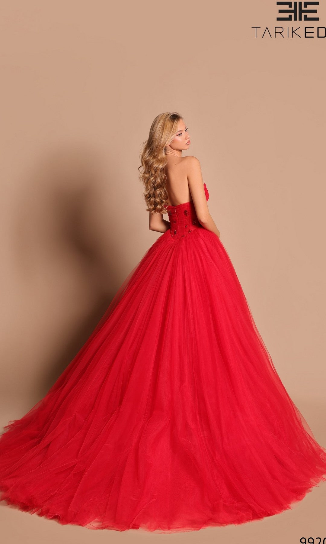 A woman with long blonde hair wears the Tarik Ediz 99205 Red Corset Ball Gown featuring a strapless design and tulle skirt, standing against a plain beige background.